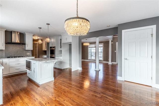 kitchen featuring a center island, backsplash, wall chimney range hood, hanging light fixtures, and ornate columns