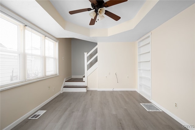 unfurnished living room featuring ceiling fan, light wood-type flooring, built in features, and a tray ceiling