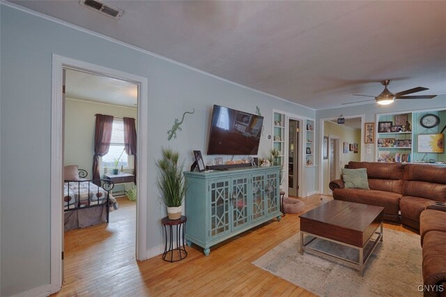 living room featuring built in shelves, ceiling fan, ornamental molding, and light wood-type flooring