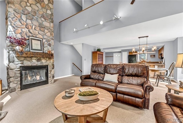 living area with baseboards, carpet, a high ceiling, a stone fireplace, and a notable chandelier