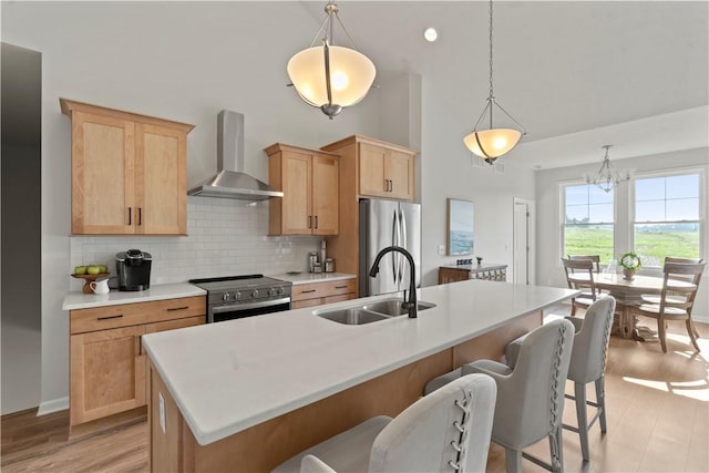 kitchen with wall chimney range hood, backsplash, decorative light fixtures, a center island with sink, and appliances with stainless steel finishes