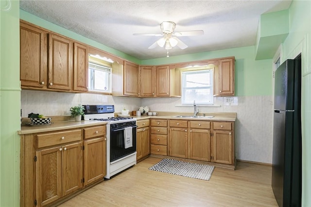 kitchen featuring white gas range, ceiling fan, sink, black fridge, and light hardwood / wood-style floors