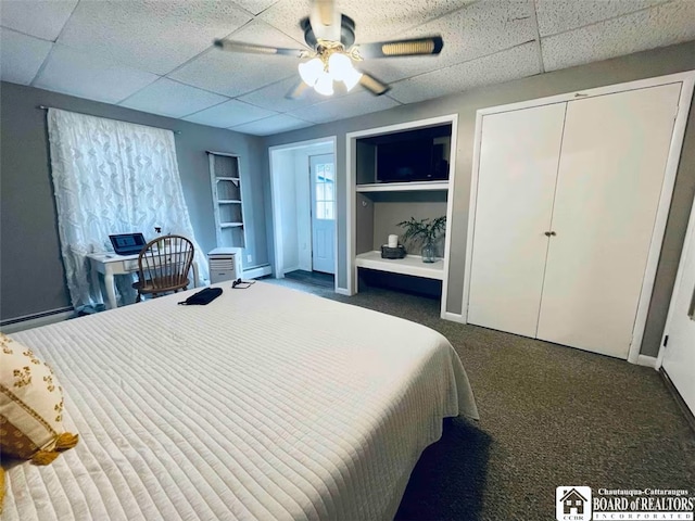 bedroom featuring a drop ceiling, dark colored carpet, ceiling fan, baseboard heating, and a closet