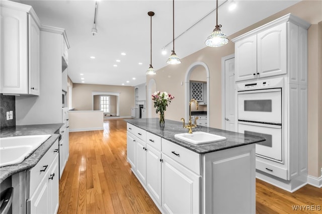 kitchen with dark stone counters, white double oven, a kitchen island with sink, white cabinetry, and hanging light fixtures