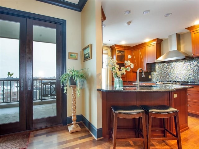 kitchen with light hardwood / wood-style floors, dark stone counters, french doors, wall chimney range hood, and tasteful backsplash