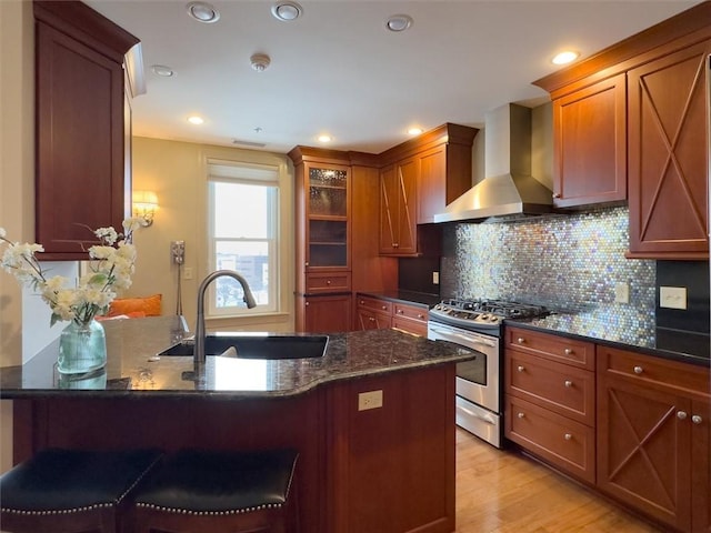 kitchen with sink, stainless steel gas stove, a kitchen breakfast bar, dark stone countertops, and wall chimney range hood