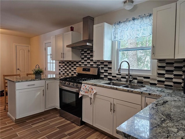 kitchen featuring white cabinetry, stainless steel gas range oven, wall chimney exhaust hood, light stone counters, and sink