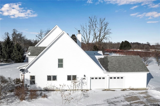 snow covered property featuring a garage, roof with shingles, and a chimney