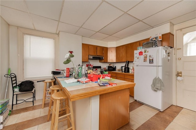 kitchen featuring a kitchen bar, kitchen peninsula, light tile patterned flooring, white refrigerator, and a drop ceiling