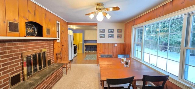 carpeted dining area featuring crown molding, wood walls, ceiling fan, and a brick fireplace