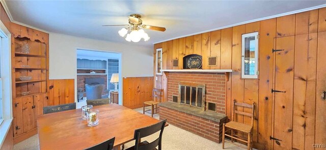 dining area featuring ceiling fan, light colored carpet, a brick fireplace, ornamental molding, and wooden walls