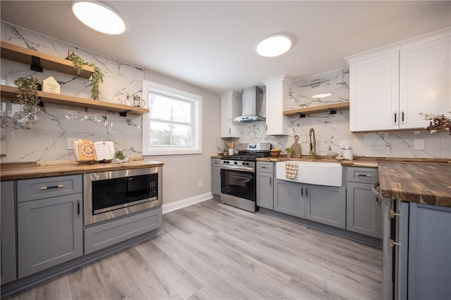 kitchen featuring built in microwave, stainless steel gas range, wooden counters, wall chimney range hood, and white cabinets