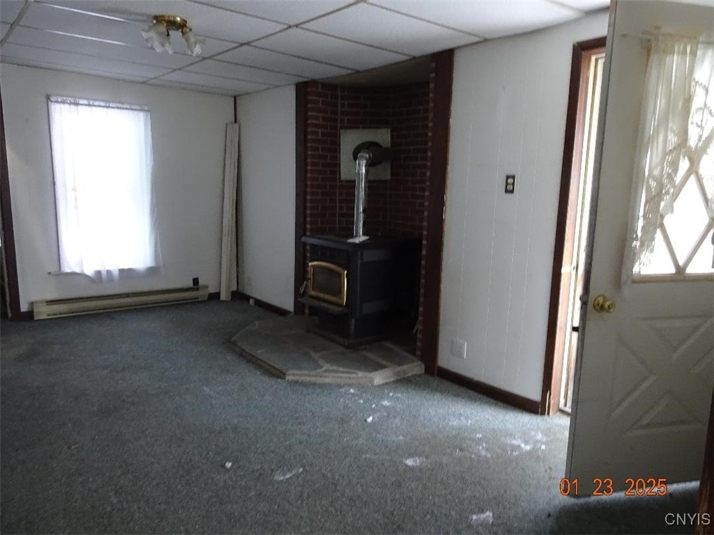 unfurnished living room with a drop ceiling, carpet floors, a wood stove, and baseboard heating