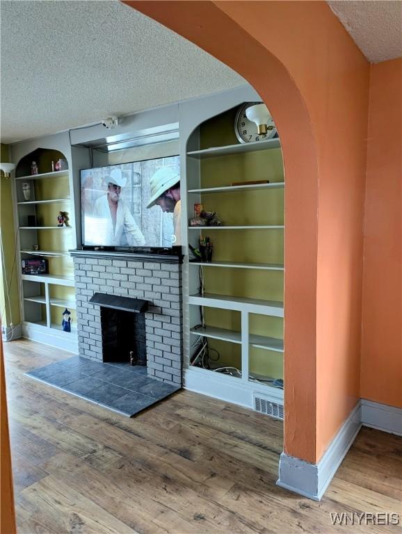 unfurnished living room with a brick fireplace, built in shelves, baseboards, wood finished floors, and a textured ceiling