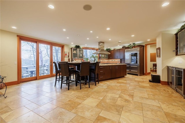 kitchen featuring island range hood, an island with sink, a breakfast bar area, stainless steel built in fridge, and dark brown cabinets