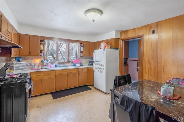 kitchen featuring sink, white refrigerator, wooden walls, stainless steel range with gas stovetop, and decorative backsplash