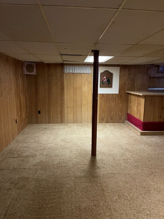 basement featuring carpet flooring, a paneled ceiling, bar area, and wooden walls