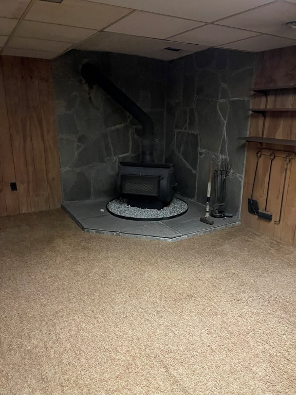 basement with carpet flooring, a paneled ceiling, wooden walls, and a wood stove