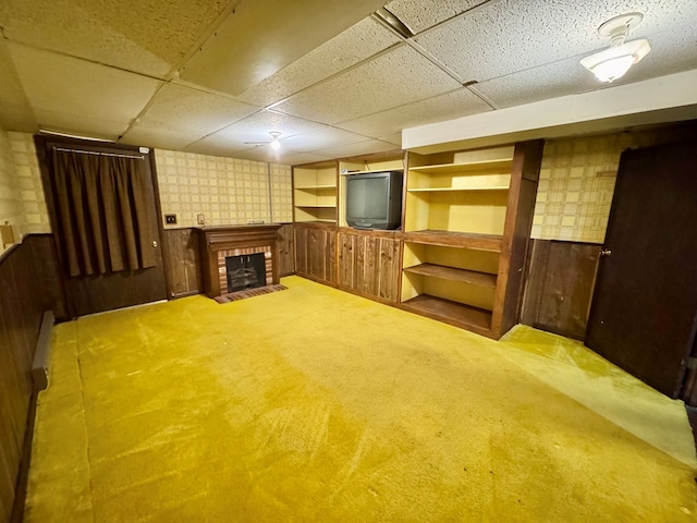 basement featuring a brick fireplace, a paneled ceiling, light carpet, and wood walls