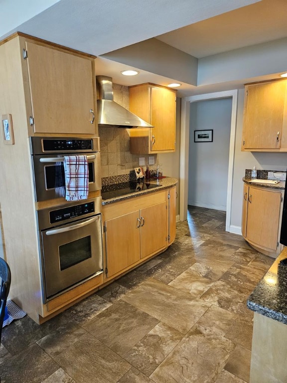 kitchen with dark stone countertops, black electric cooktop, double oven, decorative backsplash, and wall chimney range hood