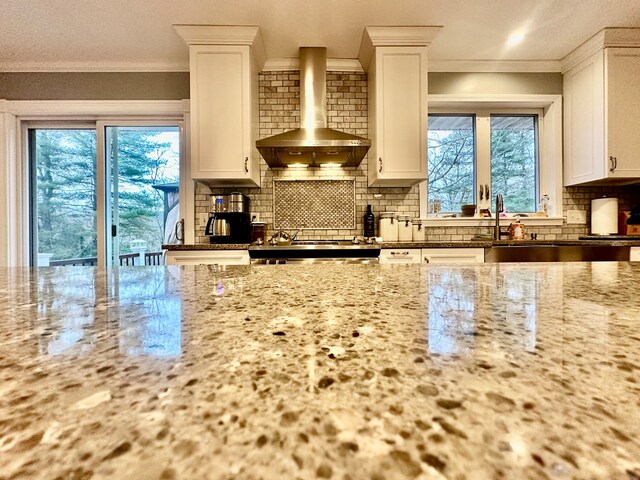 kitchen featuring wall chimney range hood, crown molding, backsplash, light stone countertops, and white cabinets