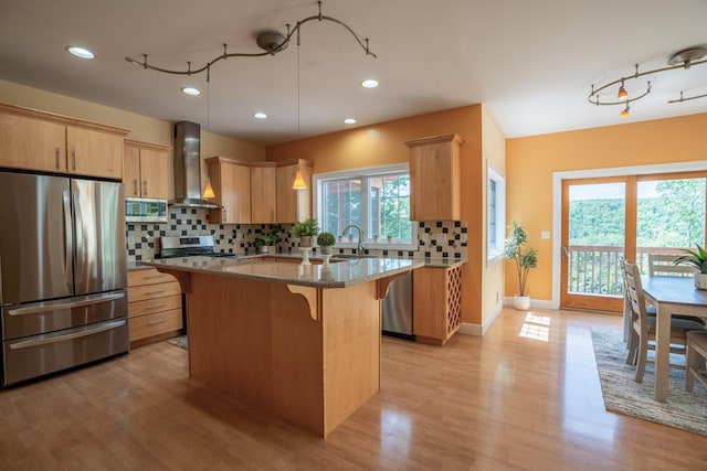 kitchen with stainless steel appliances, light stone countertops, a kitchen island, light brown cabinetry, and wall chimney exhaust hood