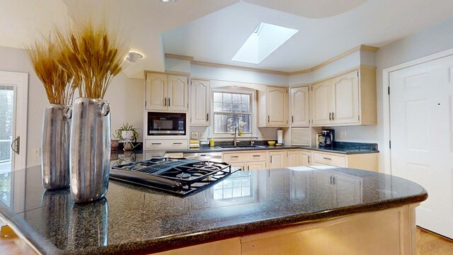 kitchen with a skylight, built in microwave, sink, dark stone counters, and cream cabinets