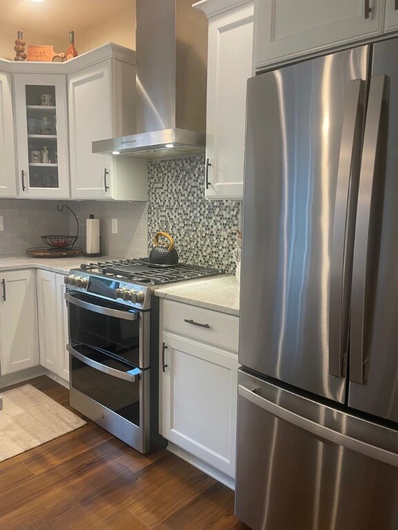 kitchen with dark wood-type flooring, wall chimney exhaust hood, white cabinetry, tasteful backsplash, and stainless steel appliances