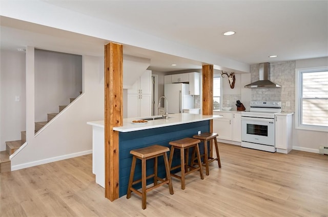 kitchen with wall chimney range hood, white appliances, sink, backsplash, and white cabinets