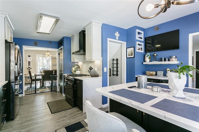 kitchen featuring stainless steel refrigerator, range with gas cooktop, white cabinets, a chandelier, and wall chimney exhaust hood