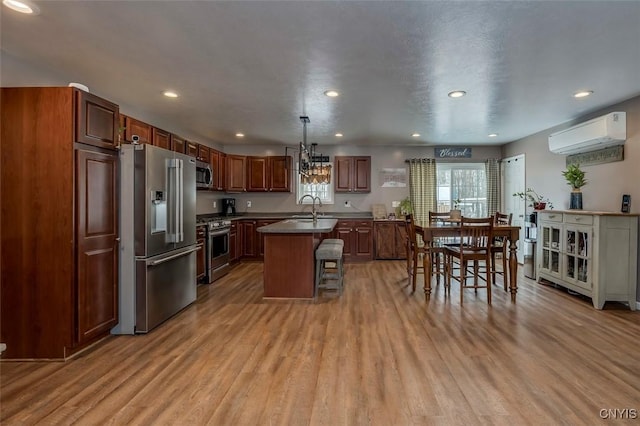 kitchen featuring appliances with stainless steel finishes, a wall unit AC, light hardwood / wood-style floors, a center island with sink, and decorative light fixtures