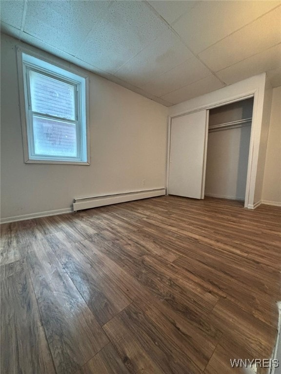 unfurnished bedroom with a drop ceiling, a baseboard heating unit, a closet, and dark hardwood / wood-style floors