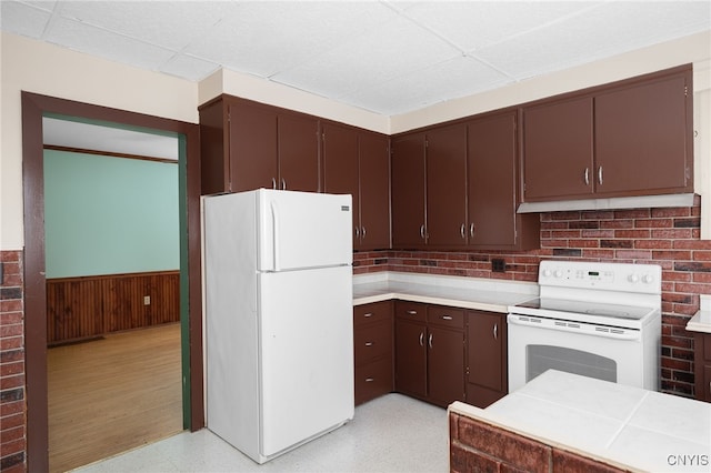 kitchen with white appliances, dark brown cabinetry, wooden walls, and a paneled ceiling