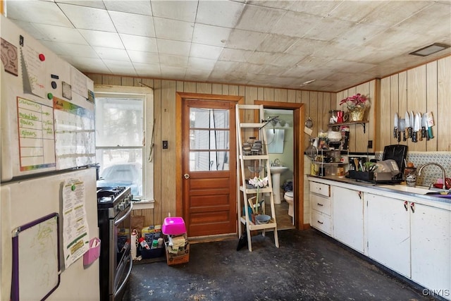 kitchen featuring sink, stainless steel gas range oven, white refrigerator, wooden walls, and white cabinets