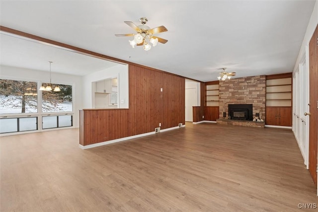 unfurnished living room featuring built in shelves, wood walls, a stone fireplace, wood finished floors, and ceiling fan with notable chandelier