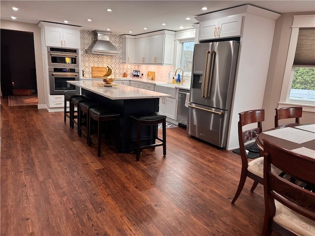 kitchen featuring appliances with stainless steel finishes, white cabinetry, dark hardwood / wood-style flooring, a kitchen breakfast bar, and wall chimney range hood