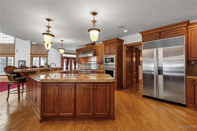 kitchen with light hardwood / wood-style flooring, built in appliances, a center island with sink, decorative light fixtures, and wall chimney exhaust hood