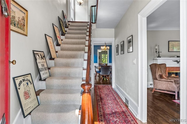 stairway with wood-type flooring, a brick fireplace, ornamental molding, and a notable chandelier