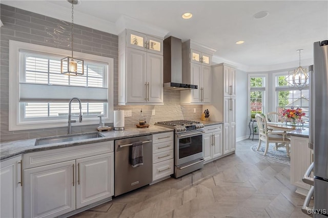 kitchen featuring appliances with stainless steel finishes, sink, wall chimney range hood, white cabinets, and hanging light fixtures