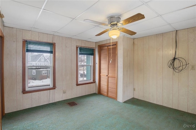 carpeted empty room with ceiling fan, a paneled ceiling, and wood walls