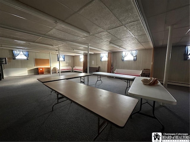 rec room with a paneled ceiling, wood walls, a healthy amount of sunlight, and baseboard heating