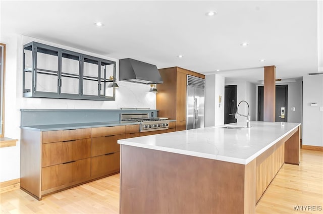 kitchen featuring appliances with stainless steel finishes, sink, a large island with sink, light hardwood / wood-style floors, and wall chimney exhaust hood