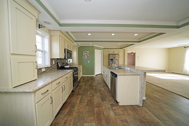 kitchen with appliances with stainless steel finishes, dark hardwood / wood-style flooring, a raised ceiling, an island with sink, and cream cabinetry