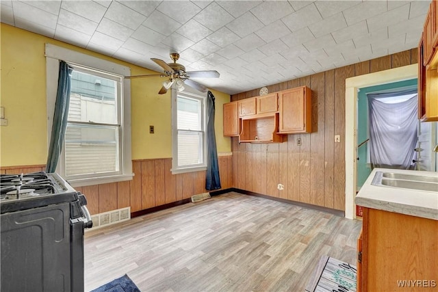 kitchen featuring sink, wooden walls, ceiling fan, gas range oven, and light hardwood / wood-style floors