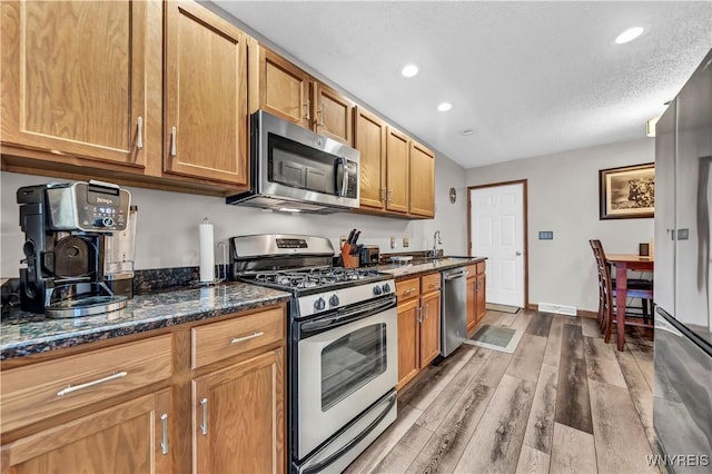 kitchen with sink, a textured ceiling, light wood-type flooring, dark stone countertops, and appliances with stainless steel finishes