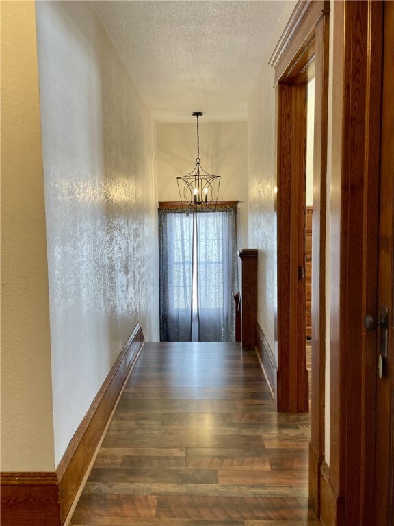 hallway featuring a textured ceiling, wood finished floors, baseboards, a chandelier, and a textured wall
