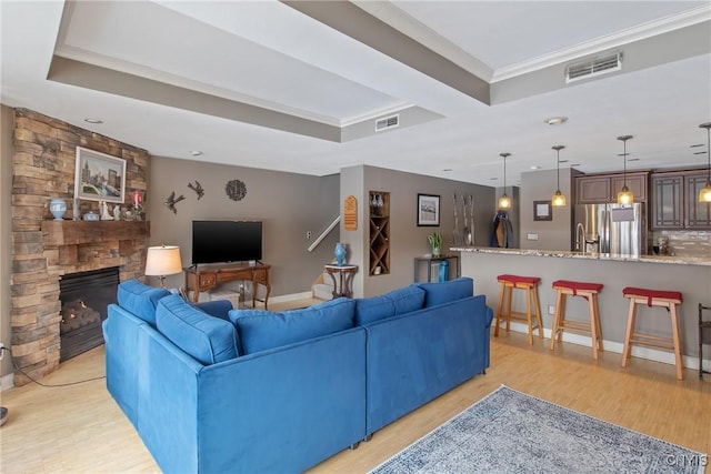 living room with ornamental molding, a tray ceiling, a stone fireplace, and light hardwood / wood-style floors