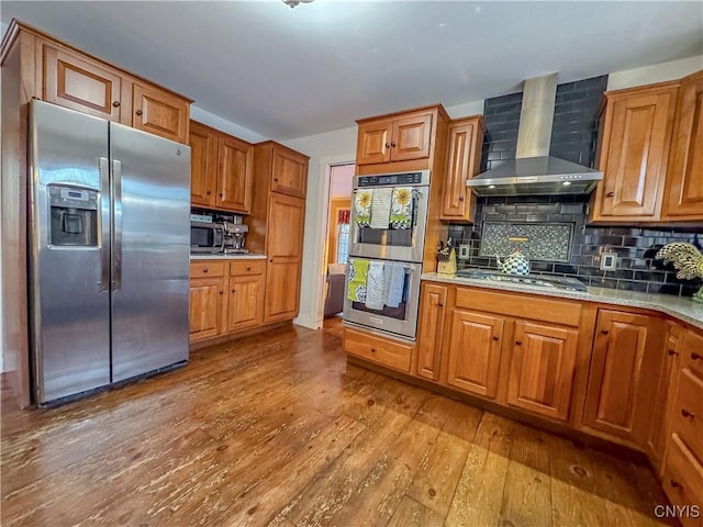 kitchen featuring wall chimney range hood, stainless steel appliances, light stone counters, light hardwood / wood-style floors, and decorative backsplash