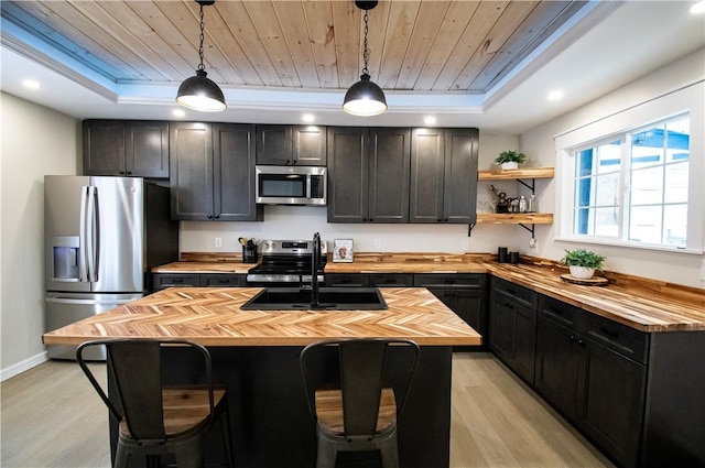 kitchen with a raised ceiling, appliances with stainless steel finishes, a center island with sink, and butcher block countertops