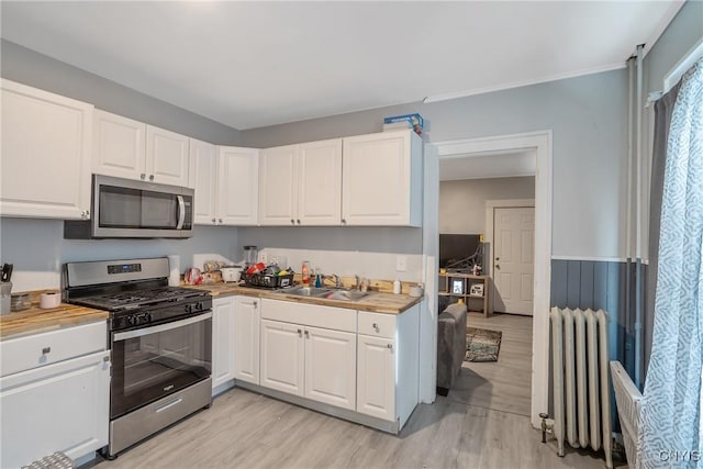 kitchen with appliances with stainless steel finishes, radiator heating unit, white cabinetry, sink, and light hardwood / wood-style floors